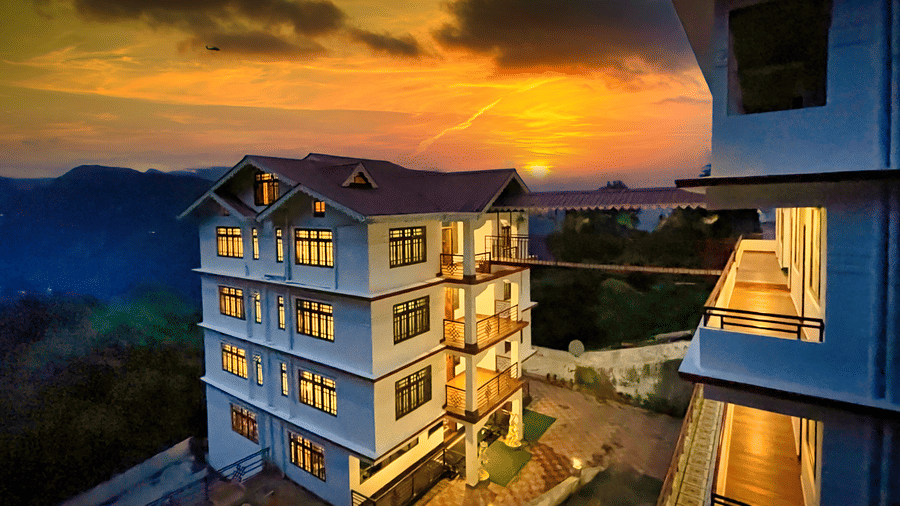 Voyage The Royal Retreat, a hillside hotel illuminated with warm yellow lights at night, set against a dramatic deep blue cloudy sky with a paved driveway and mountain silhouettes surrounding the property.