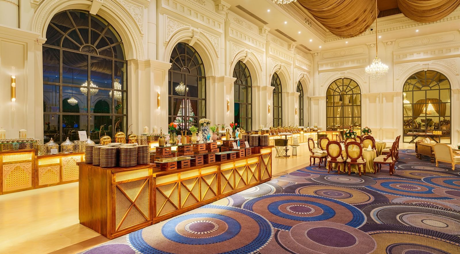 A grand dining hall at Nirvana Luxury Hotel, Ludhiana, featuring ornate architectural details, large arched windows, a buffet spread, and elegantly set tables on a patterned carpet.