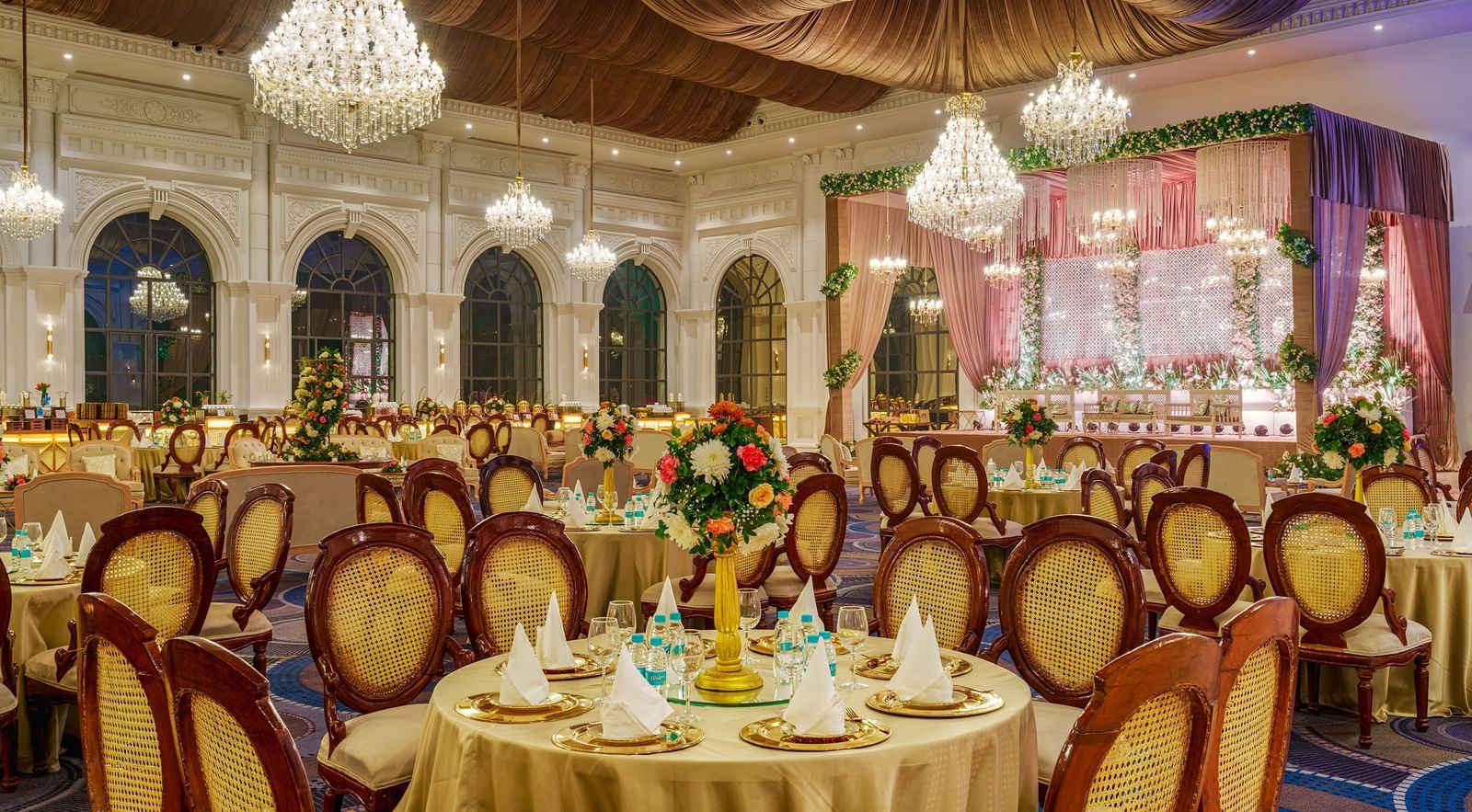  An elegant event hall at Nirvana Luxury Hotel, Ludhiana, set up for a special occasion with round tables draped in gold, ornate chairs, floral centrepieces, and sparkling chandeliers.