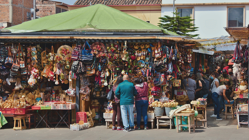 A vibrant outdoor market scene features numerous stalls selling various goods under a green canopy, with people browsing the merchandise.