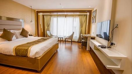 A premium hotel room with a large bed, hardwood flooring, a seating area by the large window, and a wall-mounted television, all bathed in natural light at Yashshree The Imperial Inn, Siliguri.