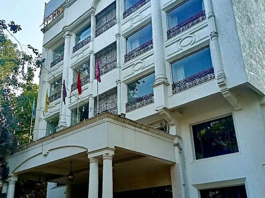  Exterior view of a multi-storey white building with large glass windows and a covered entrance supported by columns, surrounded by trees.