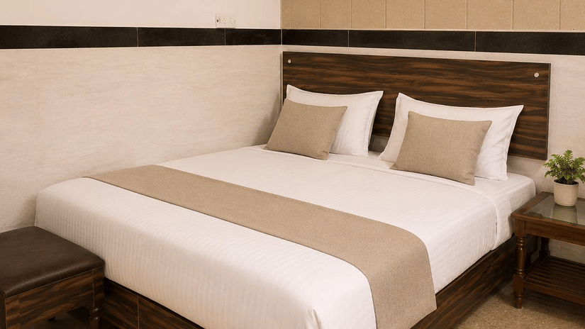 Bedroom interior of Hotel Goverdhan, Agra, featuring king size bed, white mattress cover, runner, 2 pillows, wooden headboard, side table, potted plant, stool, wall light, and tile border.