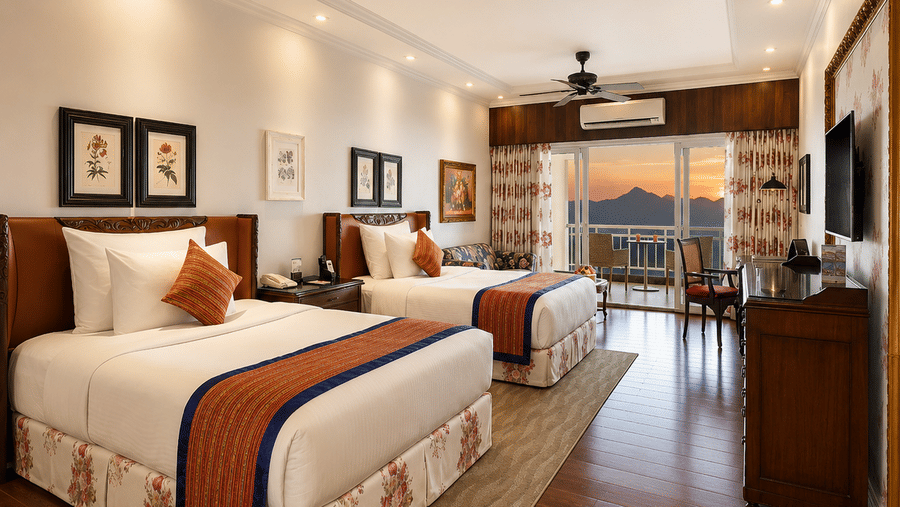 Deluxe Sunset (with Balcony) room featuring two beds placed side by side in a room at MAYFAIR Himalayan Spa Resort Kalimpong. Ceiling fans hang from the ceiling. A television rests on a console table. A balcony door is visible at the end of the room.