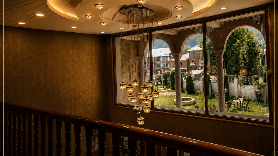 A high-angle view of a hallway at Hotel Outlook, Pahalgam, with a wooden bannister, a hanging light fixture, and arched windows showing the outdoors.