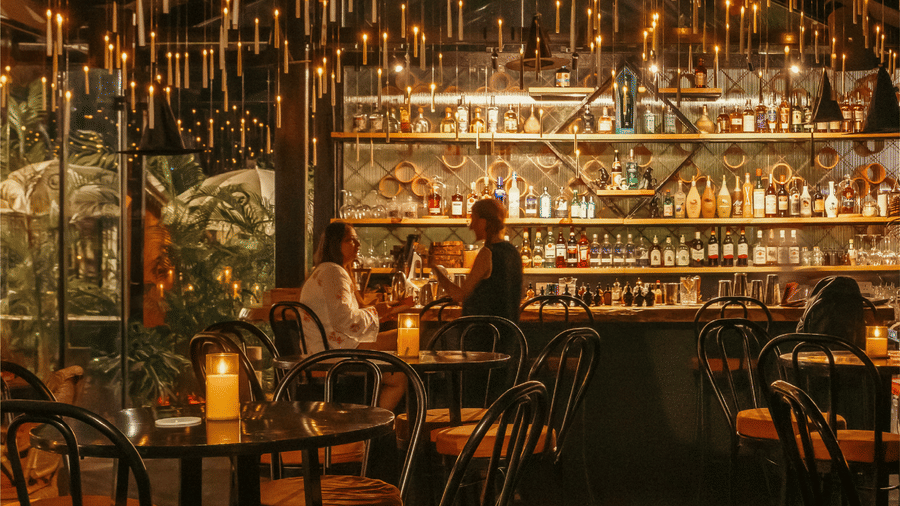 A warmly lit bar interior at The Spirit Bar featuring a backlit liquor shelf guests seated at the counter hanging pendant lights dark furniture and lush greenery in a moody atmospheric setting.