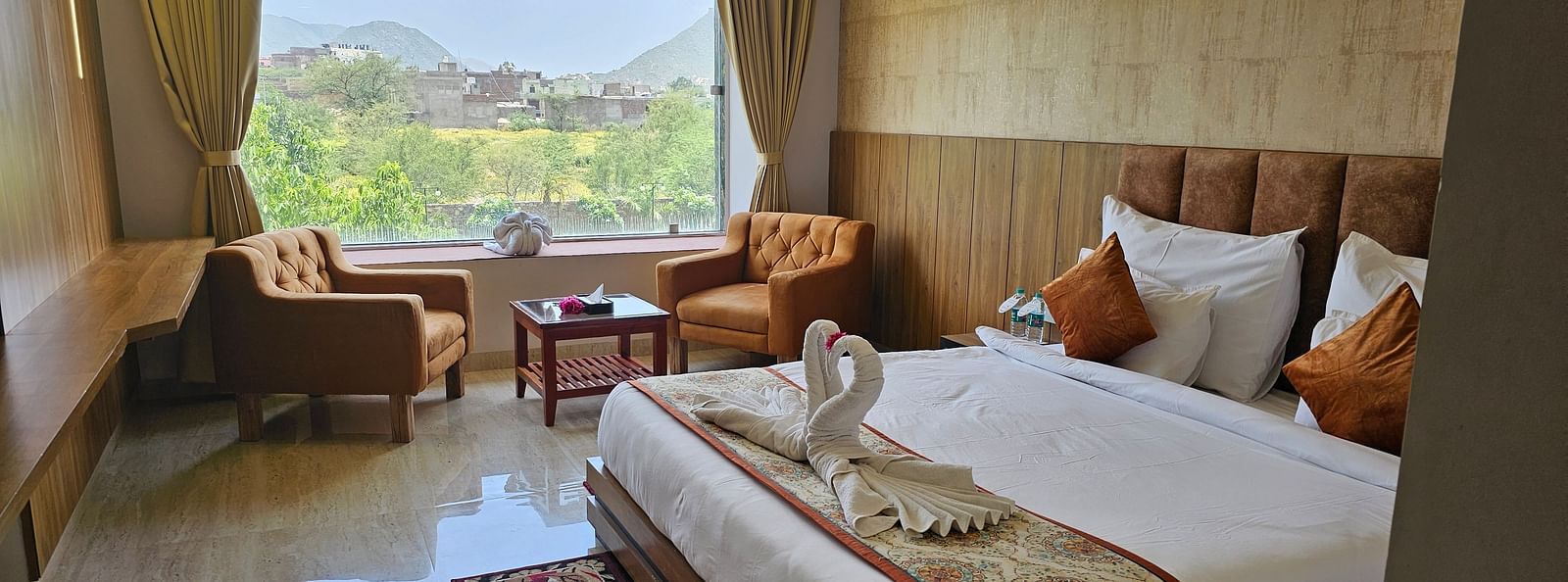 A bright and spacious resort room at Bamboo Saa Mulberry Resort, Pushkar, featuring a large window with a view of the outdoors including a made bed with a patterned runner, two armchairs, and a small table.