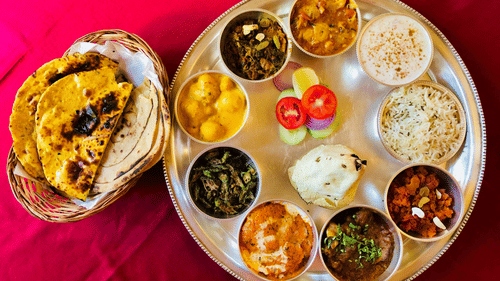 A large, round metal platter (thali) containing several small bowls (katoris) of Indian food, including curries, rice, yogurt, and a side of flatbread, placed on a red tablecloth