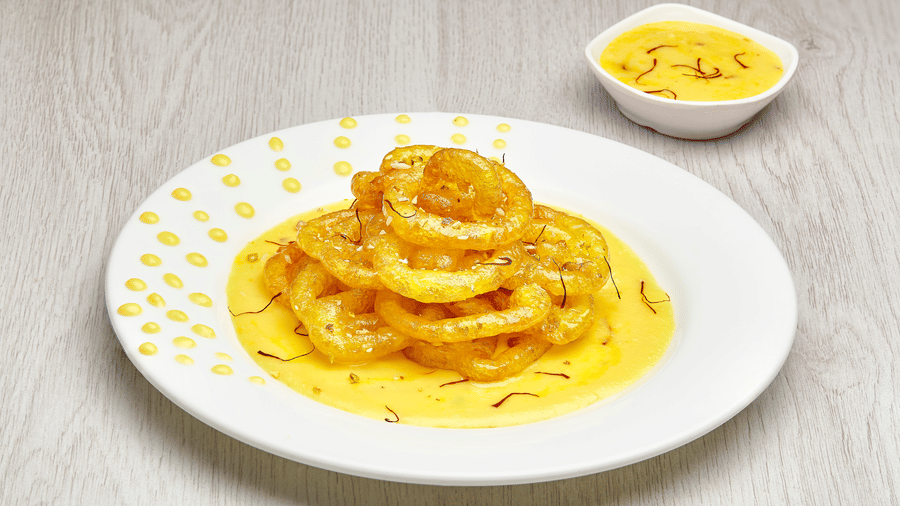 White plate of classic jalebi coils soaked in saffron rabri, accompanied by a small bowl of additional saffron rabri on a wooden table.
