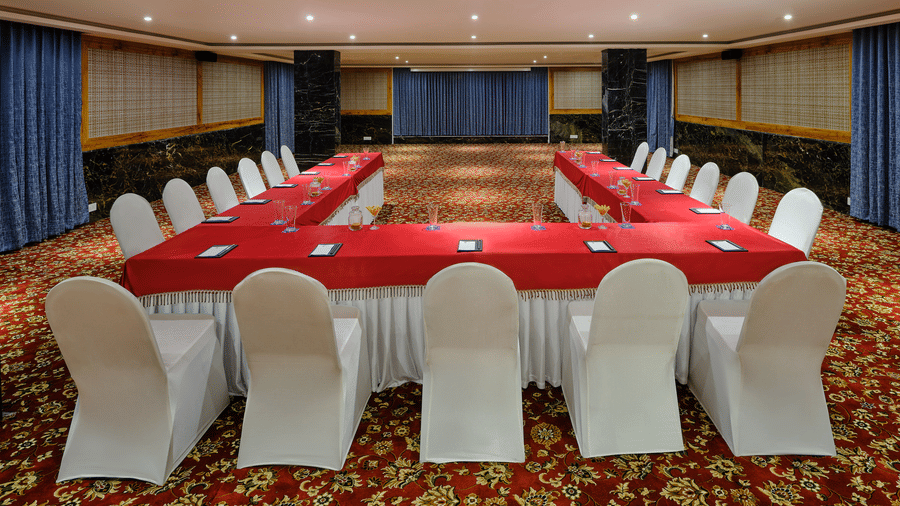 A well-organised conference room at Essentia Premier Hotel, the best hotel in OMR Chennai featuring U-shaped tables draped in red, white chairs, and a patterned carpet.