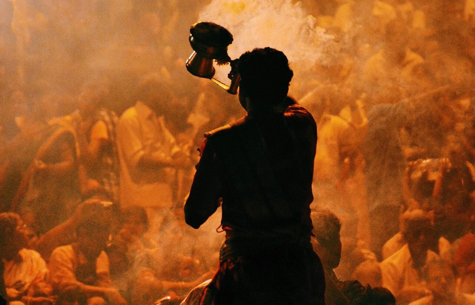 A close-up of a person performing Ganga Aarti, holding a lamp, surrounded by an engaged crowd. The scene captures the spiritual atmosphere