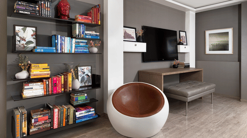 A contemporary living area at The Atara, Gurugram featuring a tall, dark bookshelf filled with books, a wall-mounted television above a sleek desk, and a distinctive circular armchair