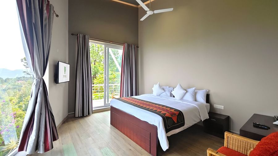 Bedroom with a bed and curtains drawn open, revealing a view of trees through glass doors at Ibex Resorts, Coonoor (Tapas).