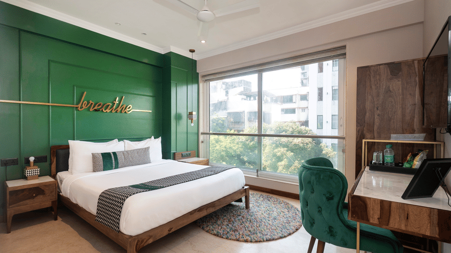 A bedroom at Theory9 - Premium Service Apartments, Bandra, featuring a large bed with white linens and a green accent wall with the word 'breathe' in gold, a wooden nightstand, a desk with a green velvet chair, and a large window overlooking trees.