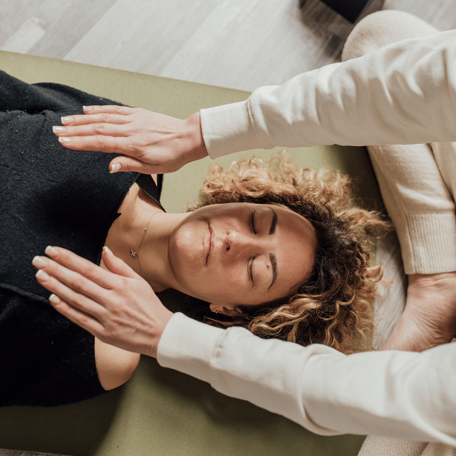 Woman lying on a yoga mat receiving energy healing or Reiki therapy, as a practitioner hovers hands above her body in a calming setting