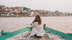 A woman sitting on a boat looking at the ghats and cityscape of Varanasi across the Ganga River.