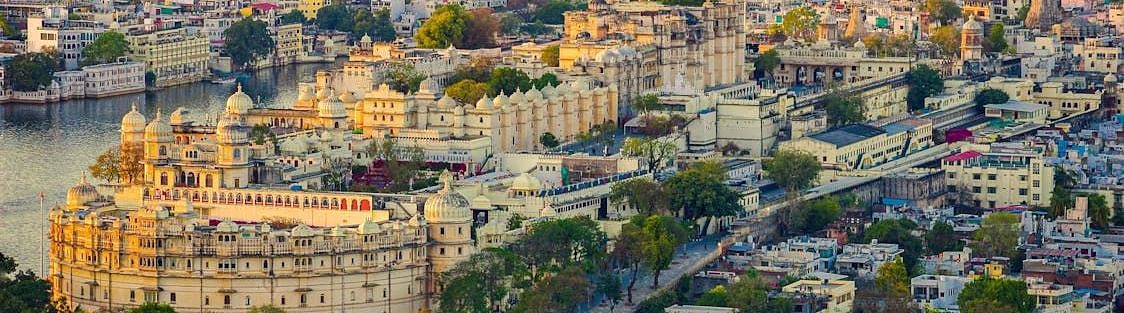 An aerial view of the city of Udaipur with the City Palace, other buildings, a lake and a hill in the background.