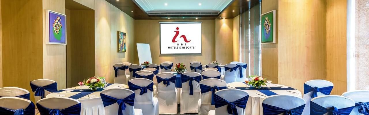 Meeting room at Inde Reva Resorts Rishhikesh, with white chairs tied with navy blue sashes, round tables decorated with flower arrangements, and a projector screen at the front.