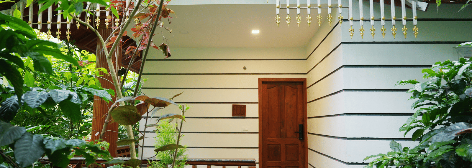 The exterior of a white, wooden-clad resort chalet with a small porch, surrounded by lush, tropical green foliage at Coorg Orange Blossom Resort & Spa.