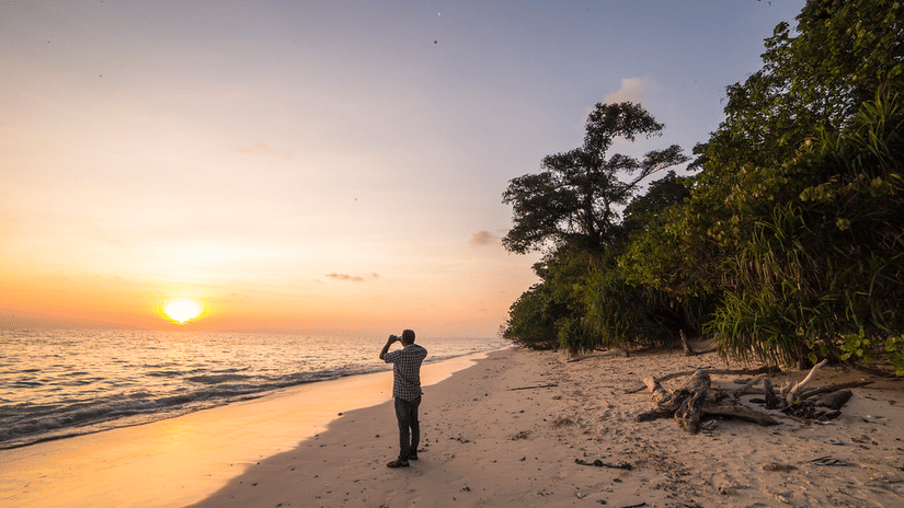 A person taking a picture of the rising sun at Kalapathar Beach while standing on the shoreline and trees next to him.