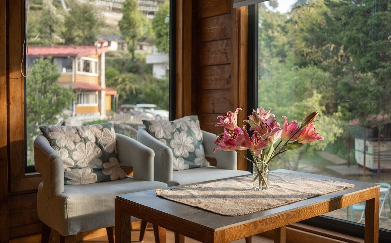 An image of sofa chairs with table near a glass wall from where sunlight is flooding the room | Avalon Cottages, Kanatal