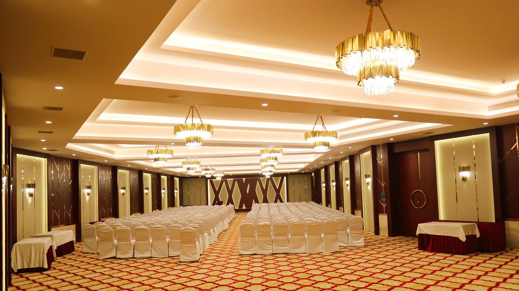 Spacious, elegant banquet hall at Jenneys Residency with patterned carpet, rows of chairs covered in cloth, decorative chandeliers, and warm lighting, set up for an event or conference