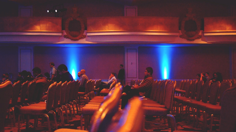 Dimly lit theatre interior with rows of seats, several people seated, and blue spotlight beams against the back wall.