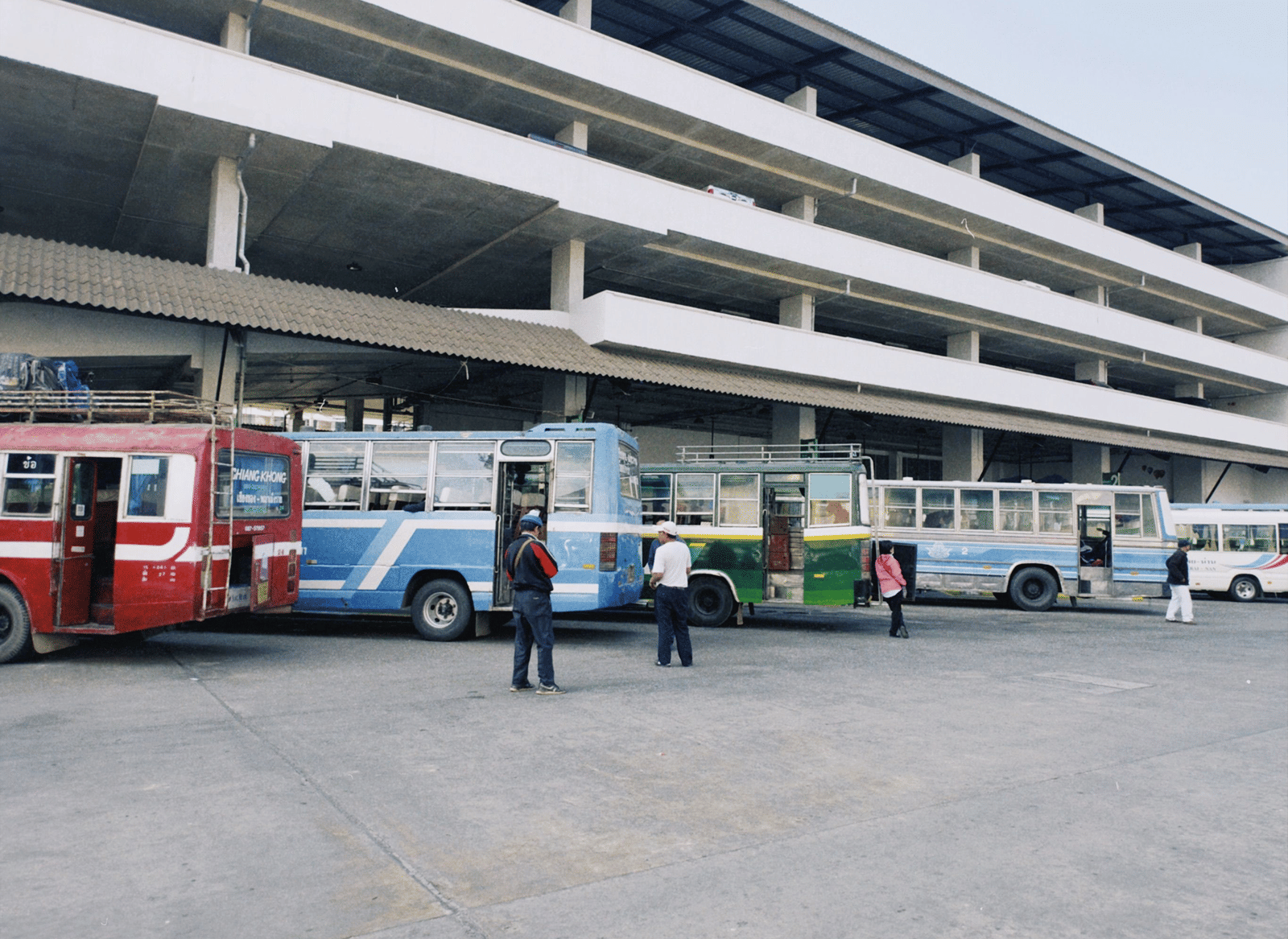 Row of 4 red, blue, green, and white buses parked at a bus stand beside a multi-storey concrete building, with a few passengers waiting on the forecourt.