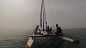 A small sailboat with multiple people aboard sails on calm water under a bright, hazy sky.