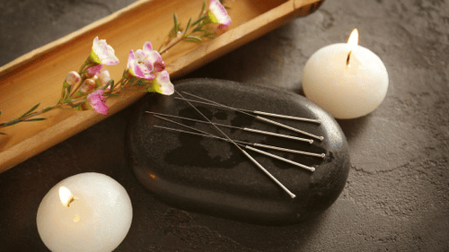 Acupuncture needles resting on a stone, next to a bamboo tray with flowers and two lit round candles