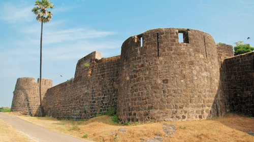 A stone fort wall with round bastions and a pathway with a single coconut tree beside it.