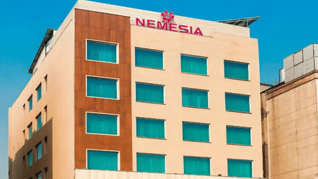 Daytime view of the Nemesia hotel building facade with teal-tinted windows, wooden accents, and palm trees in front.