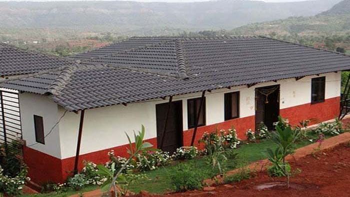 A long, single-story building with a dark roof, white upper walls, and a red lower section, situated on a hillside with a valley in the background.