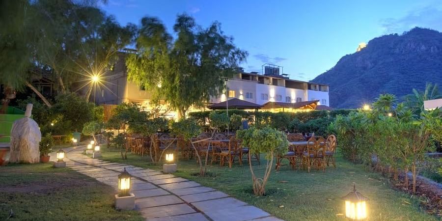 An outdoor scene at dusk where a pathway leads toward a building, which is illuminated with warm lights - Bamboo Saa Resort & Spa, Udaipur