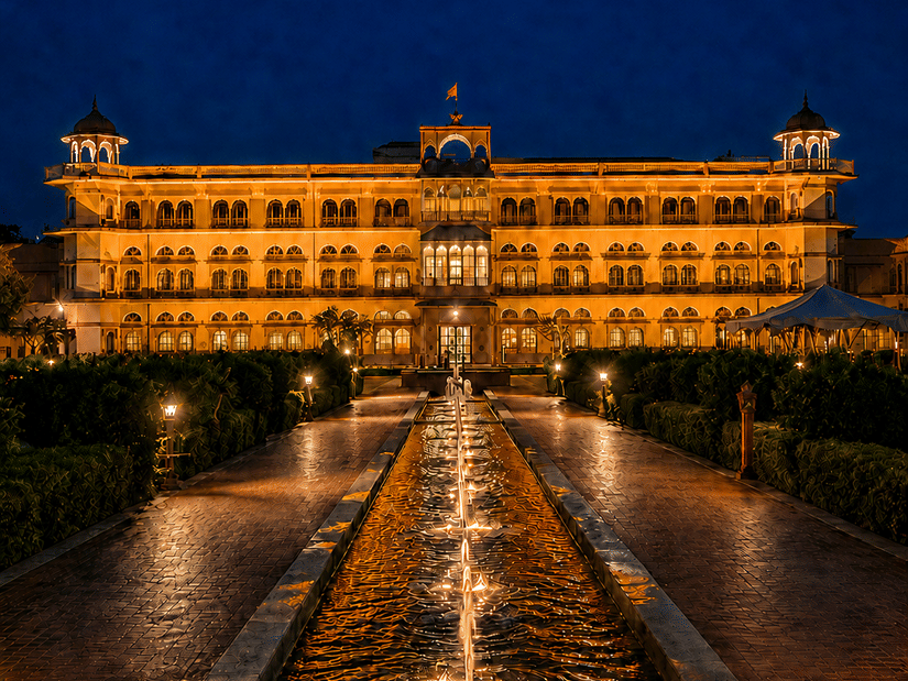 Umaid Palace glowing under a dark blue night sky, with a long reflecting pool and central walkway leading towards the building.