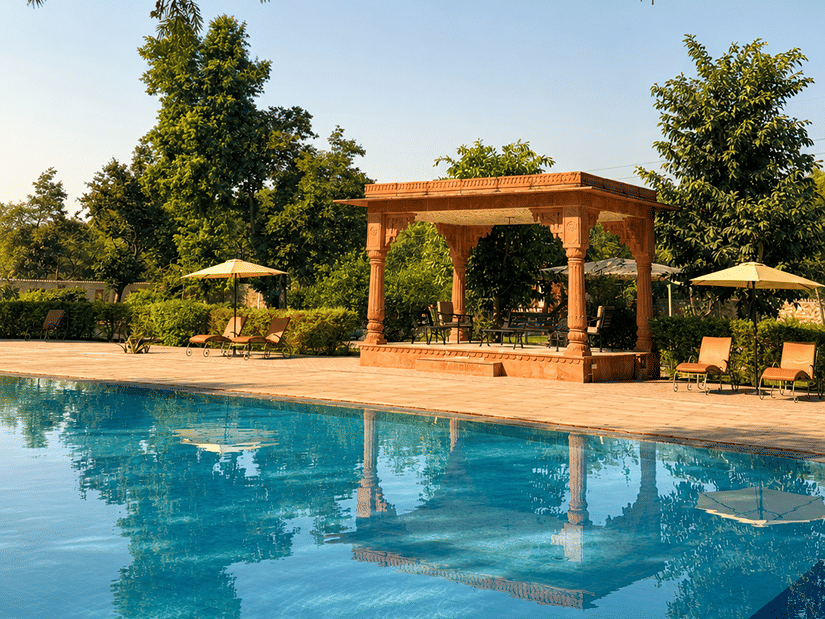 The blue swimming pool at Umaid Palace, bordered by terracotta paving, leafy trees and a sandstone pavilion.