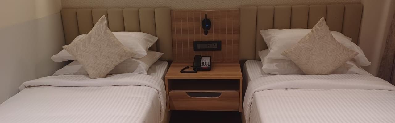 Premium Room at Hotel Paai, Hosur featuring twin beds placed at two corners of the room. Above each bed there are lights on the wall. Between the beds there is a bedside table with a telephone and socket. 