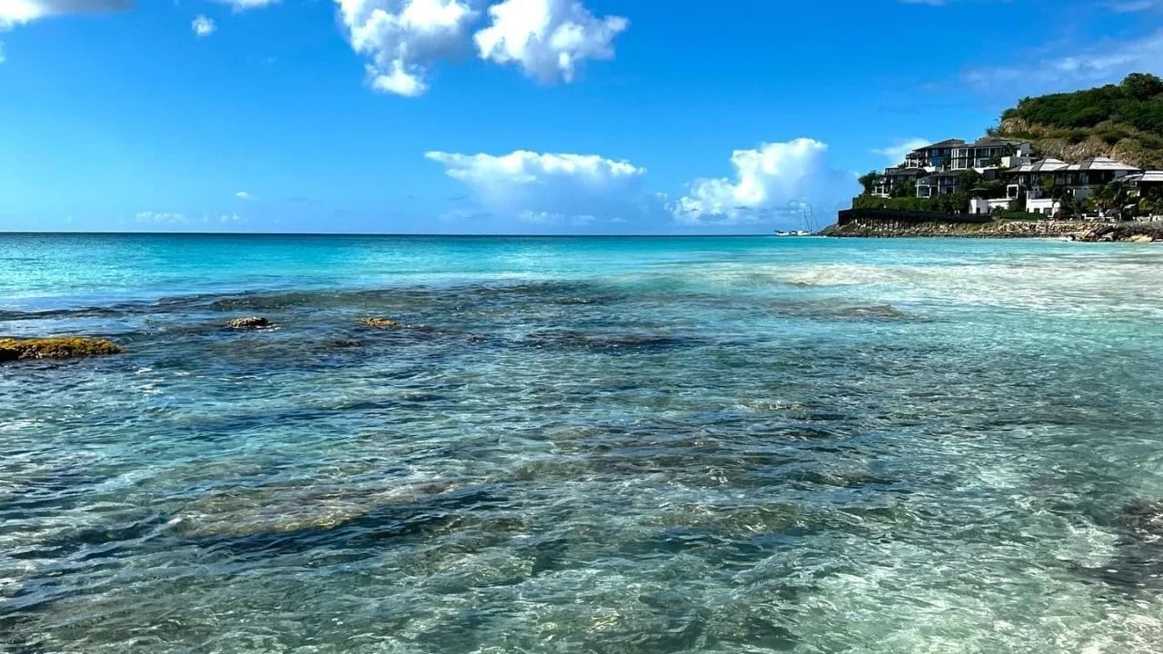 Crystal-clear turquoise ocean water with visible rocks gives way to a white sandy beach under a blue sky.