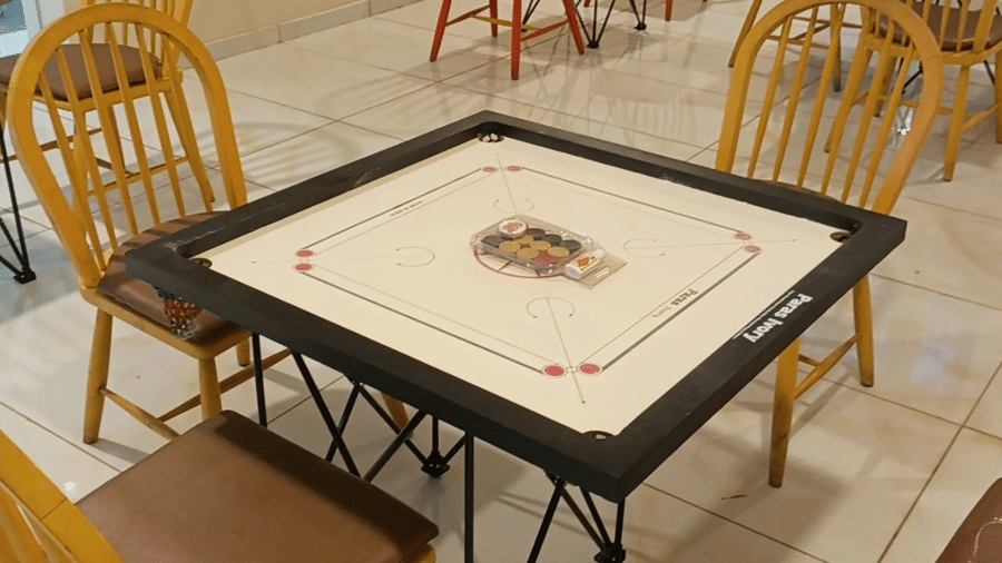 A carrom board game is set up for guests in the indoor games room at Vinti Resort, Ambaji, surrounded by wooden chairs.