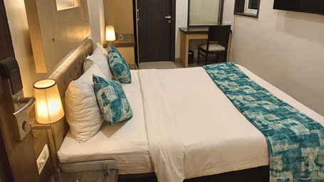 Standard Room at Hotel Mumbai House, Thane, featuring a neatly made bed with teal patterned pillows and bed runner, side lamps, wall-mounted TV, and a work desk.