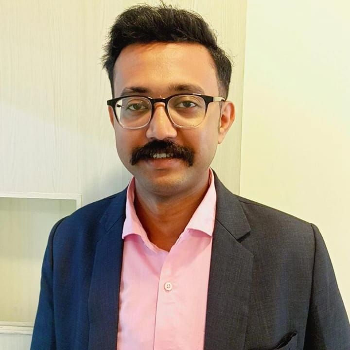 A shot of a man wearing a black blazer and salmon coloured shirt, posing in front of a cream coloured wall | Vedic Village Spa Resort