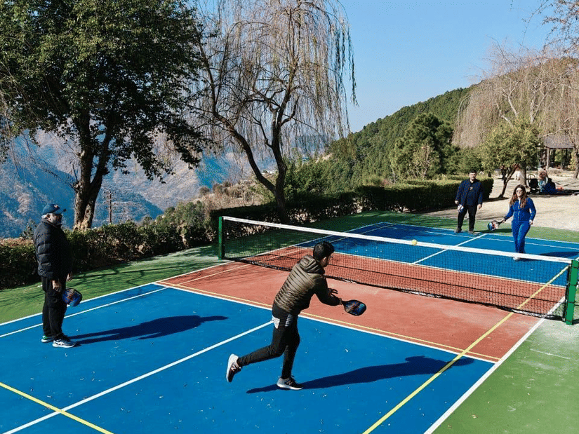 Guests playing pickle ball on vibrant blue and red court surrounded by mountain scenery and lush gardens at The Manor Wellness & Sports Hotel