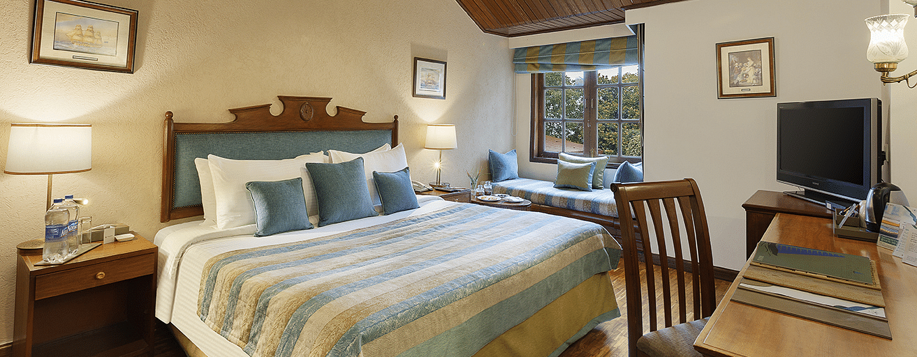 An image of a  cozy room in the naini retreat, nainital, with a king-size bed, wooden furniture, and a bay window overlooking a garden. The room features a ceiling fan, a television, and a small sitting area. 