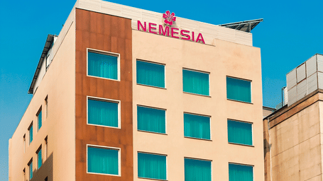 An angular view of the Nemesia City Center Hotel against a blue sky with the entrance being surrounded by trees.