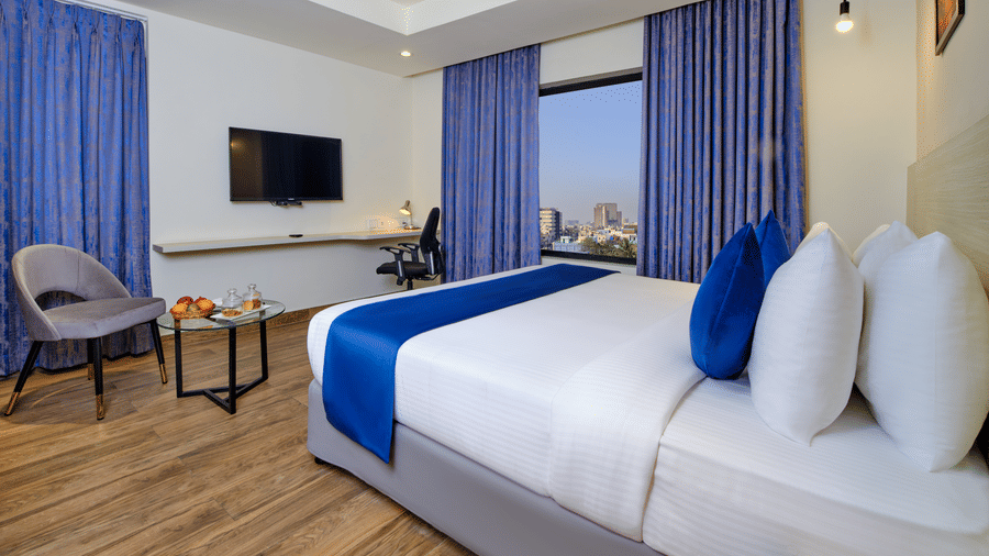 A spacious room with a king size bed set against the wall, wooden flooring beneath, a TV mounted opposite, and a cosy seating area by the window, offering a refined stay in Chennai at Essentia Premier Hotel.