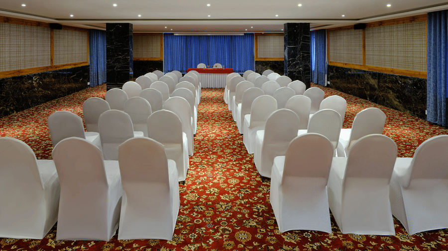 A large hall with rows of white chairs, a patterned carpet and a blue backdrop, well set up for events at meeting halls in Chennai - Essentia Premier Hotel, Chennai