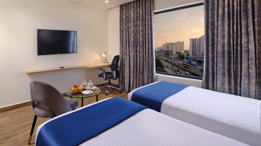 A twin-bed room with wooden flooring facing a wall-mounted TV, a seating area by the window with city views, highlighting places to stay in Chennai - Essentia Premier Hotel, Chennai