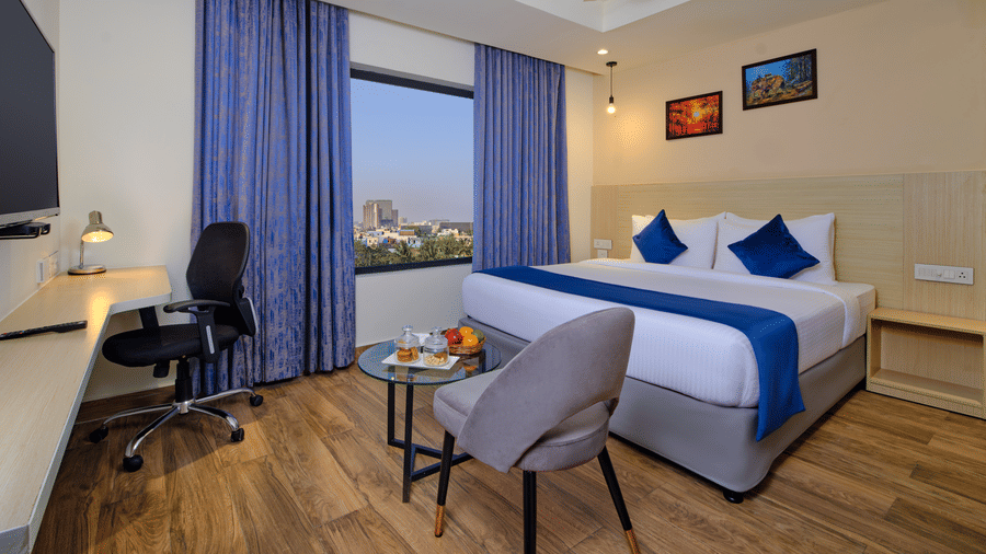 A modern bedroom with a large bed, blue accents, a work desk, and city views reflecting the comfort of our hotel rooms in Chennai - Essentia Premier Hotel, Chennai