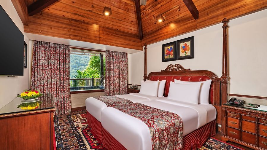 Imperial Villa bedroom with a large bed and decorative rug, a table, wooden cabinet beneath a TV , wooden ceiling and a window with decorative curtains overlooking greenery