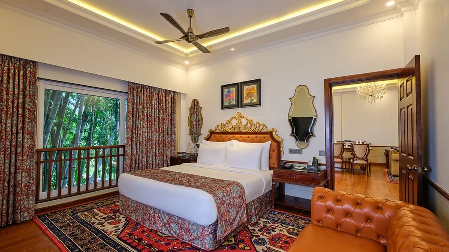 a beautiful Suite with a bed and decorative rug, sofa, mirror, artworks and a huge window with curtains overlooking greenery 